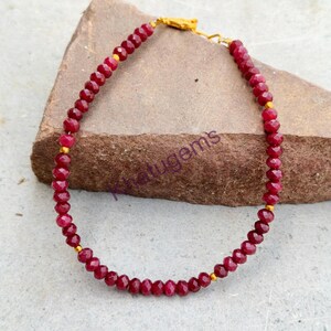 Op de afbeelding: Een kralenarmband met kleine, gefacetteerde, dieprode edelsteenkralen in een cirkelvormig patroon, afgewisseld met kleine, gladde, goudkleurige metalen tussenkralen en een goudkleurige metalen sluiting aan de bovenzijde.