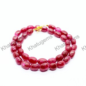 May include: A necklace composed of smooth, oval-shaped, deep red gemstone beads arranged in a circular pattern. The strand features a gold-toned metal clasp and is overlaid with the repeating watermark text Khatugems in black.