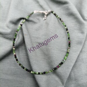 May include: A beaded necklace featuring small, faceted green and dark red stones arranged in a circular pattern. The necklace includes a silver-toned metal lobster claw clasp and a short extender chain. The text Khatugems is visible in the center.