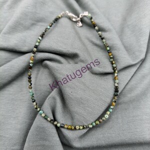 May include: A beaded necklace featuring small, round, multi-colored stones in shades of turquoise, green, brown, and black. The necklace is secured with a silver-toned metal lobster claw clasp. The text Khatugems is overlaid in purple across the center.