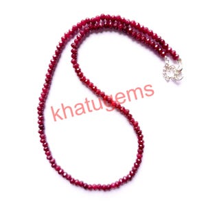 May include: A necklace composed of small, faceted, deep red gemstones. The beads are strung together, forming a delicate strand. A silver-colored clasp secures the necklace. The text "khatugems" is visible in the image.