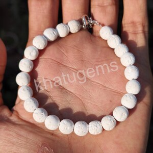 Op de afbeelding: Witte kralenarmband met een zilveren sluiting. De armband is gemaakt van ronde, getextureerde kralen. De tekst "khatugems" is zichtbaar op de armband. Deze sieraden zijn perfect voor dagelijks gebruik.