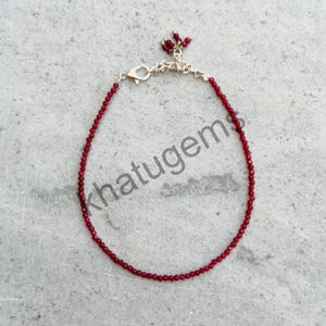 May include: A delicate red beaded bracelet with a gold-colored clasp and a small cluster of red beads at the end. The bracelet is displayed on a gray surface. The text "khatugems" is visible on the image.