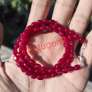 Puede incluir: Un collar compuesto por piedras preciosas ovaladas de color rojo intenso. Las cuentas están ensartadas, formando un bucle continuo. Un cierre plateado asegura el collar. La palabra "hatugem" es parcialmente visible.