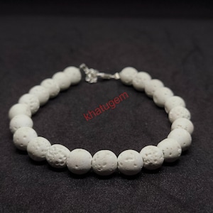 Puede incluir: Una pulsera de cuentas blancas con un cierre plateado. La pulsera está hecha de cuentas redondas y texturizadas. La palabra "khatugem" está impresa en rojo en la imagen. La pulsera se muestra sobre un fondo oscuro.