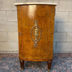 May include: An antique wooden cabinet with a marble top. The cabinet has a warm, brown finish with a decorative brass oval featuring a classical figure. The cabinet is supported by four short legs. The cabinet is approximately 76 cm tall.