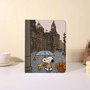 Könnte beinhalten: Eine Tablet-Hülle mit Snoopy-Motiv, die Snoopy und Woodstock zeigt, die im Regen unter einem Regenschirm spazieren gehen. Der Hintergrund ist eine detaillierte Illustration einer Stadtstraße mit Gebäuden und einem Uhrturm. Die Hülle ist beige und hat ein glänzendes Finish.