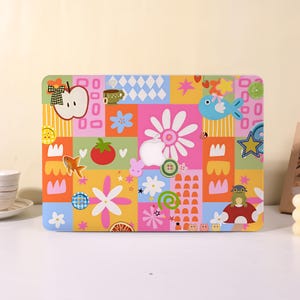 May include: A laptop cover featuring a vibrant, multi-colored patchwork design with various illustrations including an apple, a fish, a tomato, a mushroom, a frog, flowers, stars, buttons, and a cup, centered around a white Apple logo cutout.