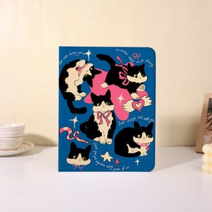 May include: A blue rectangular notebook cover featuring a repeating pattern of black and white cats with pink bows and collars. The design includes a large pink star, small white stars, hearts, and cursive text that reads "I'm not leaving you," "no matter what we go through," and "I'm always with you."