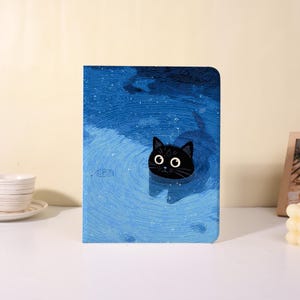 May include: A blue notebook with a whimsical illustration of a black cat swimming in water. The cat has large, round eyes and a curious expression. The background features a textured, watery design.