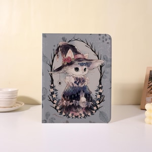 Pode incluir: Uma capa cinza para tablet com uma ilustração fantasiosa de um gato vestido de bruxa. O gato usa um chapéu preto com rosas cor-de-rosa e um vestido azul e branco. Uma aranha pende do chapéu, e o gato é emoldurado por uma grinalda de galhos e flores.
