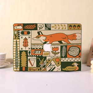 May include: A laptop hard shell case featuring a grid of illustrated nature motifs including a fox, a duck, pine trees, mushrooms, acorns, and a tree stump. The design uses a palette of orange, dark green, and cream with a central Apple logo cutout.