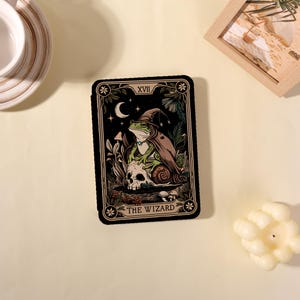 May include: A tarot card featuring a frog wizard with a crescent moon and the text "THE WIZARD". The card has a black background with botanical and celestial elements. The frog is wearing a wizard hat and cape.