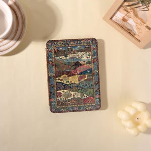 May include: A rectangular notebook featuring a detailed, colorful landscape illustration of trees, hills, and wildlife. The cover is framed by an intricate, dark blue and gold patterned border. The design includes shades of pink, green, and blue.