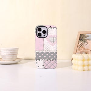 May include: A phone case featuring a patchwork design with light pink, white, and gray sections. The pattern includes small hearts, stars, plaid, and polka dots, accented with black bow illustrations and a large heart shape on the upper right side.
