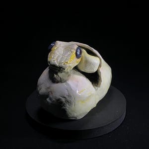 May include: A ceramic sculpture of a snake hatching from an egg. The snake has a cream-colored body with yellow accents and blue eyes. The egg is off-white with a rough texture, and the sculpture sits on a black base.