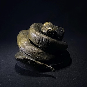 以下が含まれることがあります： ブロンズ色の巻き付いたヘビの彫刻。ヘビの頭は持ち上げられ、体はきつく巻かれています。彫刻は質感のある表面を持ち、暗い背景に配置されています。