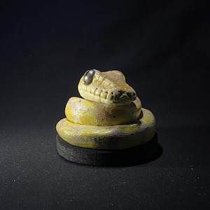 以下が含まれることがあります： 黒い目と黒い台座が付いた、黄色とタン色のコイル状のヘビの置物。ヘビの頭は持ち上げられ、体は円形のコイル状になっています。置物は暗い背景に設定されています。
