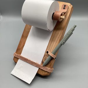 May include: A wooden notepad holder with a roll of white paper and a gray pen. The holder is made of light brown wood and features leather straps. The pen is secured with a leather loop.