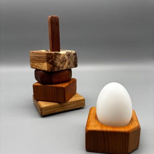 Può includere: Una pila di quattro blocchi di legno quadrati di varie tonalità di marrone con un montante verticale in legno e una base esagonale in legno separata che sostiene un uovo bianco liscio. I pezzi di legno mostrano venature e texture naturali.