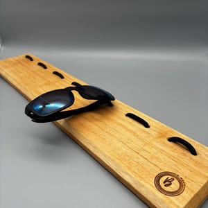 Peut inclure: Des lunettes de soleil noires avec des verres bleus reposent sur un présentoir en bois. Le présentoir est rectangulaire, avec un grain de bois clair et plusieurs élastiques noirs. Un logo circulaire est gravé dans le bois.