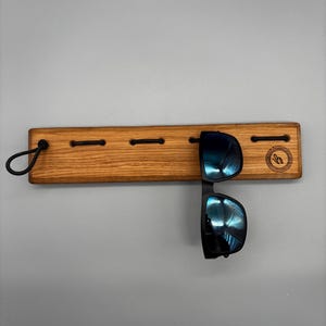 Peut inclure: Un support à lunettes de soleil en bois avec une boucle en cordon noir et quatre fentes. Le support est en bois brun clair et présente une paire de lunettes de soleil noires avec des verres réfléchissants bleus. Le support a un logo sur le côté droit.
