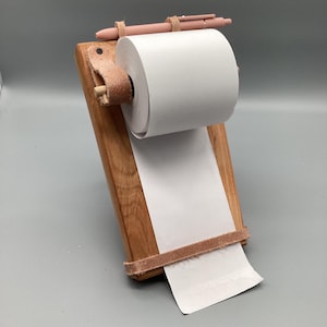 May include: Wooden notepad holder with a roll of white paper and a pink pen. The holder is made of light brown wood and has a leather strap to hold the paper in place. A pink pen rests on top of the holder.