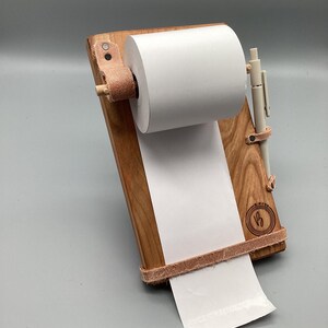 May include: Wooden notepad holder with a roll of white paper and a pen. The holder is made of light brown wood with leather accents. A pen is secured to the side with a leather strap. The paper is unrolled from the top.