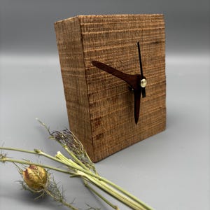 May include: A wooden block clock with dark brown hands and a brass-colored center. The clock is made of a light brown, textured wood. Dried greenery is in the foreground.