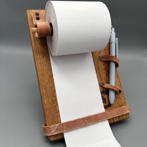 May include: A wooden notepad holder with a roll of white paper and a light blue pen. The holder has brown leather straps and a wooden dowel to hold the paper roll. The wood has a natural grain and a logo is visible.