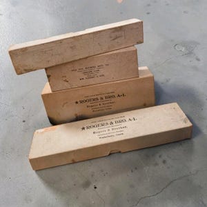 May include: A stack of four rectangular cardboard boxes in varying sizes, featuring light tan surfaces with printed black text. The boxes display branding for The Wm. Rogers Mfg. Co. and Rogers & Bro. A-1, including locations like Meriden and Waterbury, Conn.