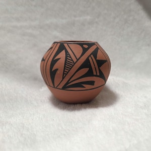 May include: A small, round terracotta-colored pottery vase with a black geometric design. The vase features a wide opening and a rounded body, showcasing a traditional Native American aesthetic.