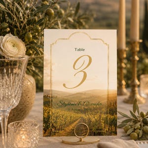 Könnte beinhalten: Eine rechteckige Tischnummernkarte mit einem Weinberg-Landschaftshintergrund und einem goldfarbenen Rand. Die Karte zeigt den Text Table 3, die Namen Stephanie & Alexander sowie das Datum 17. August 2030 auf einem runden Goldständer.
