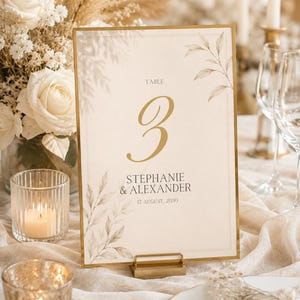 Könnte beinhalten: Ein rechteckiges Tischnummernschild mit einem goldenen Metallrahmen und Ständer. Die cremefarbene Karte zeigt den Text TABLE 3, STEPHANIE & ALEXANDER und 17 AUGUST, 2030 in dunkler Schrift, akzentuiert durch zarte, hellbraune botanische Blattillustrationen.