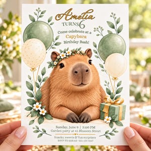 Könnte beinhalten: Eine Geburtstagseinladungskarte mit einem braunen Wasserschwein mit Gänseblümchenkranz, umgeben von grünen und cremefarbenen Luftballons, Blättern und einem Geschenk. Text: Amelia turns 6, Come celebrate at a Capybara Birthday Bash, Sunday, June 9, 3:00 PM, Garden party at 45 Blossom Street, Snacks, sweets & fun surprises, RSVP to SARAH 555-123-4567.