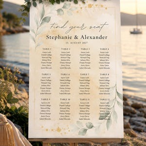 Könnte beinhalten: Ein Hochzeitssitzplan auf einer Holzstaffelei mit Aquarellhintergrund, grünen Blattillustrationen und goldenen Sprenkeln. Der Text lautet Find your seat, Stephanie & Alexander, 15. August 2027, und listet zwölf Tische auf.