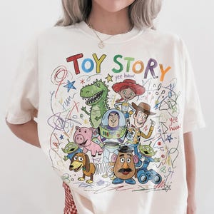 Peut inclure: T-shirt cr&egrave;me avec un dessin color&eacute; des personnages de Toy Story, dont Buzz l'&Eacute;clair et Woody. Les mots "TOY STORY" sont &eacute;crits dans une police multicolore ludique au-dessus des personnages. Le design a une esth&eacute;tique dessin&eacute;e &agrave; la main.
