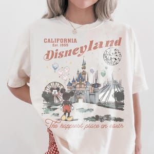 May include: Cream-colored t-shirt featuring a Disneyland graphic. The design includes the text "CALIFORNIA Disneyland Est. 1955" with an illustration of Mickey Mouse, a castle, and other park elements. The phrase "The happiest place on earth" is also included.