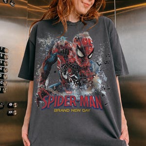 May include: A charcoal gray short-sleeve t-shirt featuring a graphic of Spider-Man in a red and blue suit. The design includes paint splatter effects, the Marvel logo, and the text "SPIDER-MAN BRAND NEW DAY" printed in bold red and yellow letters.