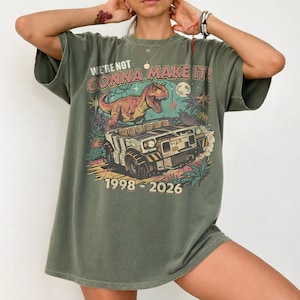 May include: An olive green short-sleeve t-shirt featuring a graphic print of a dinosaur standing on a rugged vehicle. The text reads WE'RE NOT GONNA MAKE IT! and 1998 - 2026. The design includes palm trees, stars, and a moon in a retro-inspired illustration.