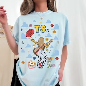 May include: A light blue t-shirt featuring a graphic of a person with blonde hair wearing a yellow plaid shirt and cow-print pants, holding a guitar and a red hat. The design includes the text TS, blue clouds, musical notes, and flowers.