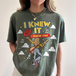 May include: An olive green t-shirt featuring a graphic print with the text I KNEW IT and I KNEW YOU. The design includes a person playing a guitar, a red hat, white clouds, yellow stars, and musical notes against the solid green fabric background.