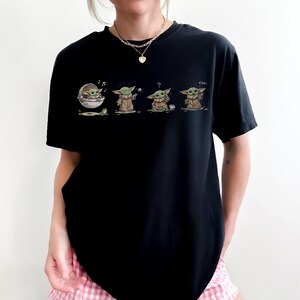 May include: A black short-sleeved t-shirt featuring a graphic print of a small green creature in four different poses. The print shows the character in a floating pod, holding a ball, looking confused with a cup, and waving with the text coo.