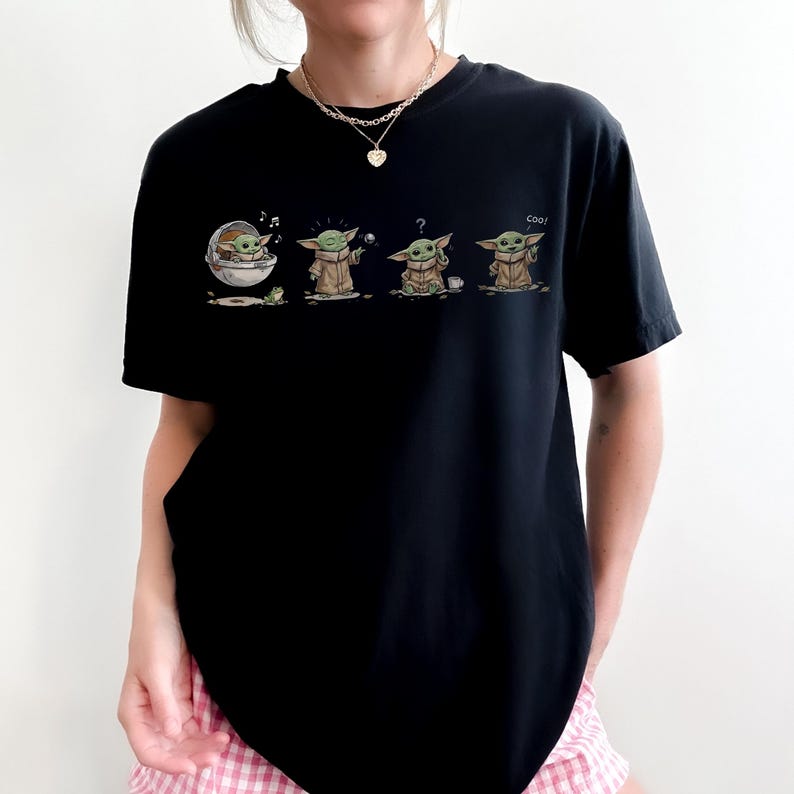 May include: A black short-sleeved t-shirt featuring a graphic print of a small green creature in four different poses. The print shows the character in a floating pod, holding a ball, looking confused with a cup, and waving with the text coo.