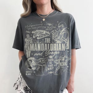 May include: A charcoal gray short-sleeve t-shirt featuring a cream-colored graphic print of The Mandalorian and Grogu. The design includes various Star Wars characters, ships, and the text: Mind your manners, You know what I'M TALKING ABOUT, THE MANDALORIAN and Grogu, THIS IS THE WAY, and WHEREVER I GO, HE GOES.