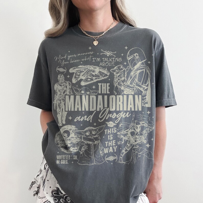 May include: A charcoal gray short-sleeve t-shirt featuring a cream-colored graphic print of The Mandalorian and Grogu. The design includes various Star Wars characters, ships, and the text: Mind your manners, You know what I'M TALKING ABOUT, THE MANDALORIAN and Grogu, THIS IS THE WAY, and WHEREVER I GO, HE GOES.