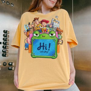 May include: A mustard yellow short-sleeve t-shirt featuring a graphic print of various Toy Story characters surrounding a green frog-shaped drawing board. The board displays the text Hi! Let's play! in blue script against a blue background.