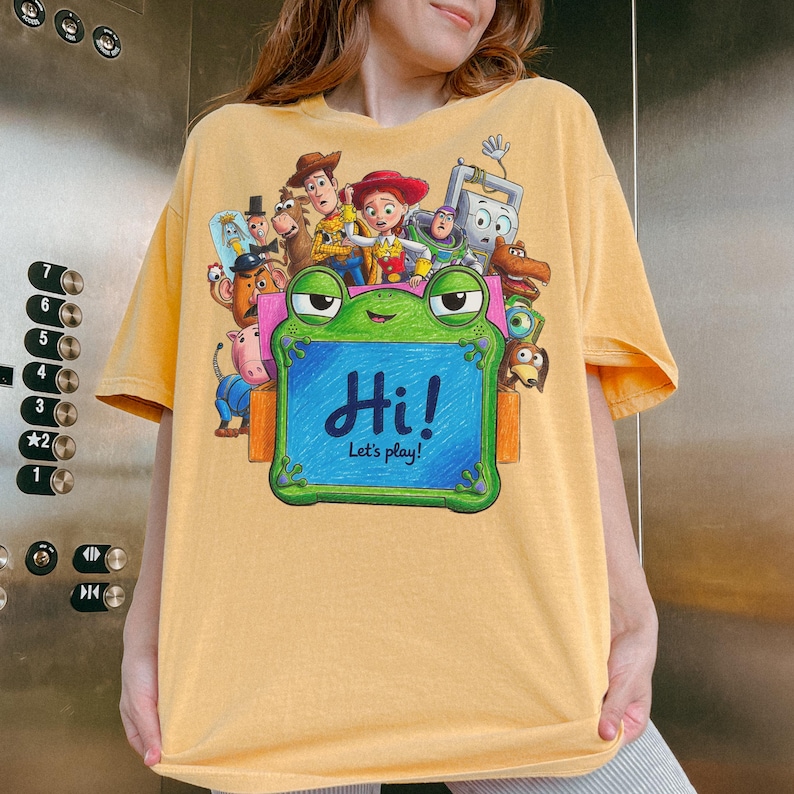 May include: A mustard yellow short-sleeve t-shirt featuring a graphic print of various Toy Story characters surrounding a green frog-shaped drawing board. The board displays the text Hi! Let's play! in blue script against a blue background.
