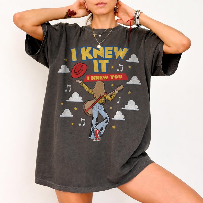 May include: A charcoal gray short-sleeve t-shirt featuring a graphic print with the text I KNEW IT and I KNEW YOU. The design includes a person with long hair playing an acoustic guitar, a red hat, musical notes, stars, and white clouds.