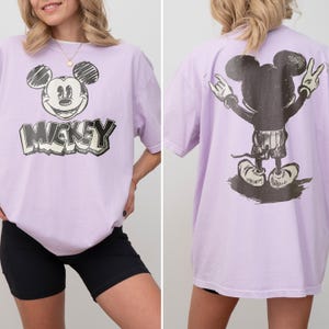 May include: A light purple short-sleeve t-shirt featuring a black and white sketch-style graphic of Mickey Mouse on the front with the text MICKEY below it, and a back graphic showing the character from behind with hands raised in a peace sign.
